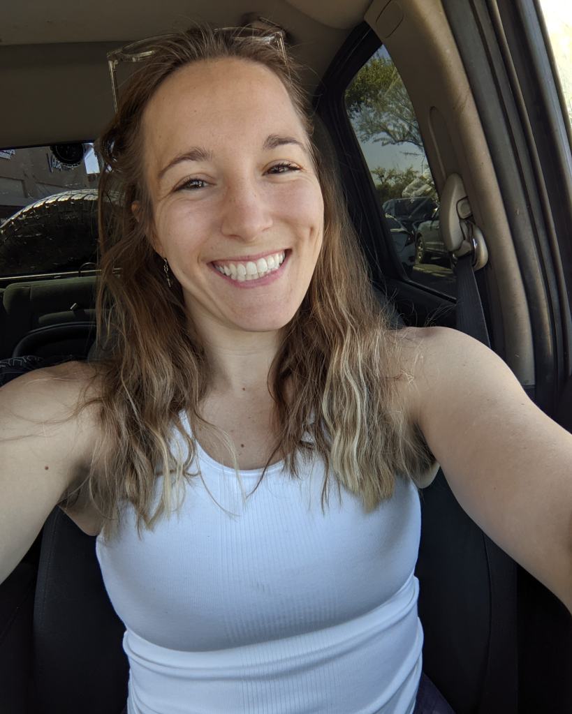 A smiling woman with long, wavy hair sitting in a car, wearing a white tank top.